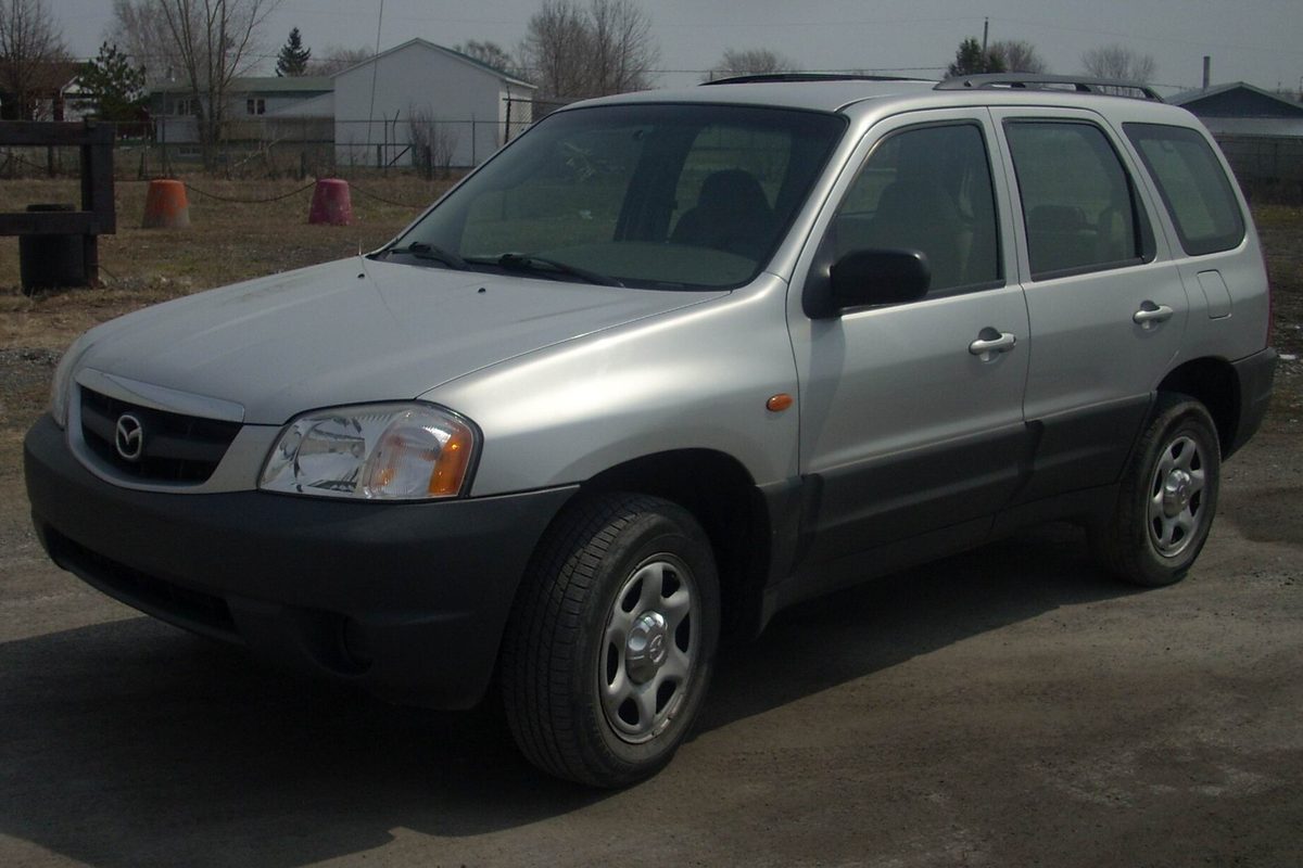 A silver 2001-2004 Mazda Tribute SUV parked on a dirt lot with bare trees and a fence behind it