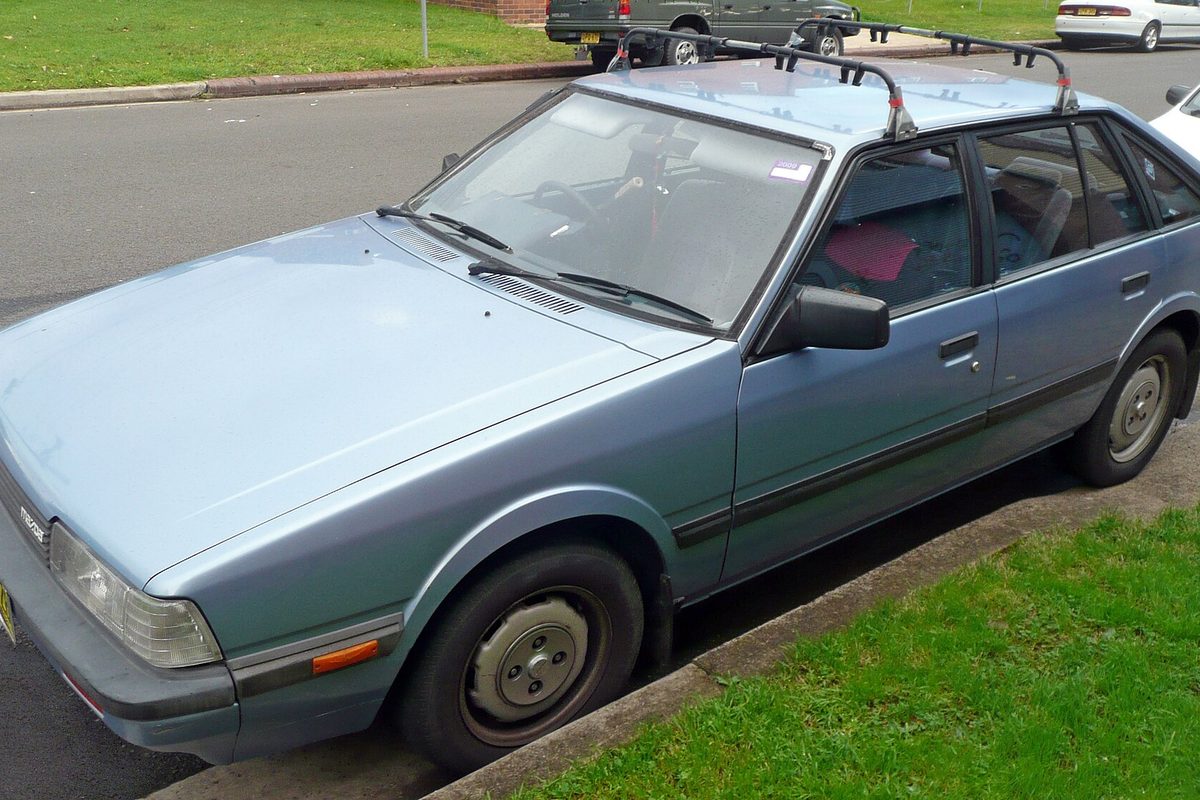 A faded blue-gray 1980s Mazda 626 hatchback parked by a grass verge on a quiet street