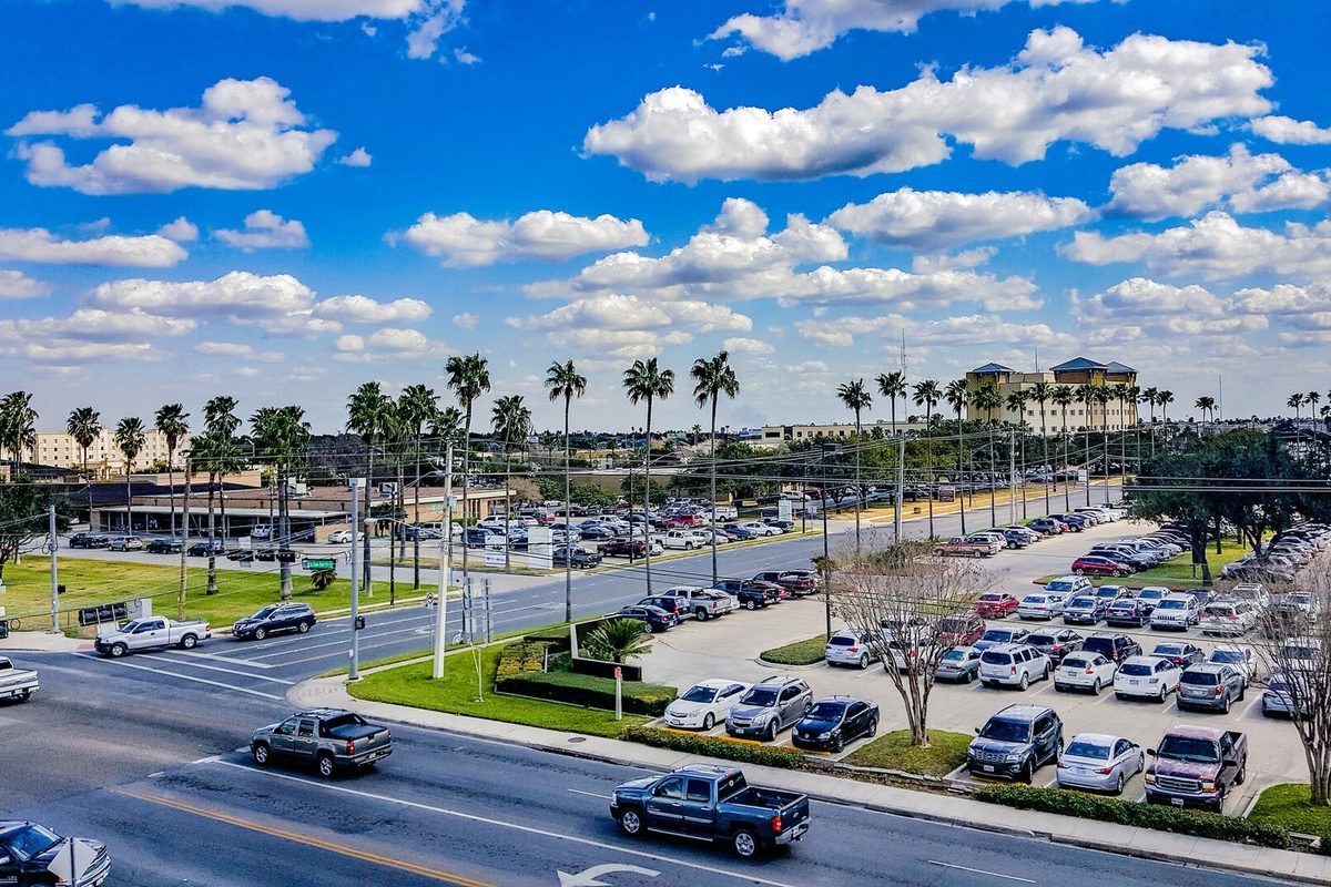 A wide McAllen street lined with tall palm trees, pickup trucks driving past a full parking lot under a blue sky with white clouds