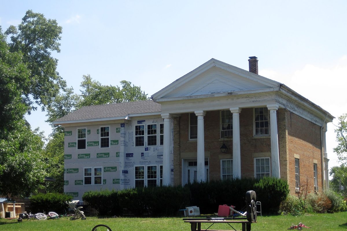 The Count's House in McHenry, a two-story tan brick house with white columns and a pediment, its side wing wrapped in house wrap during repairs