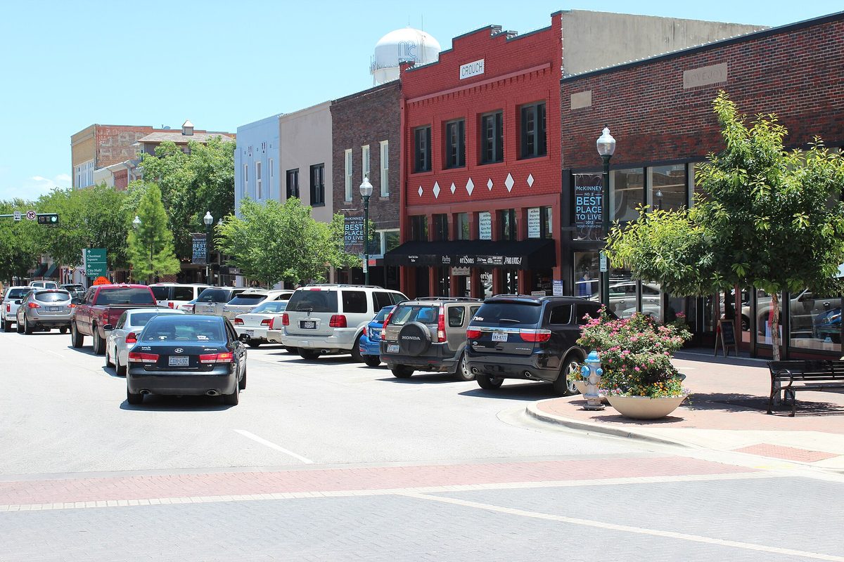 Cars parked along brick storefronts on the historic downtown square in McKinney, with a red building, street trees and a water tower behind