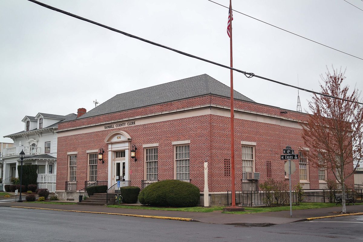 A red brick building marked Yamhill County Clerk on a McMinnville street corner, with a flagpole and rounded shrubs out front
