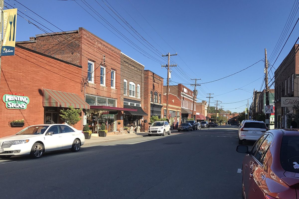 A downtown Mebane street lined with two-story brick storefronts and cars parked at the curb under a clear blue sky