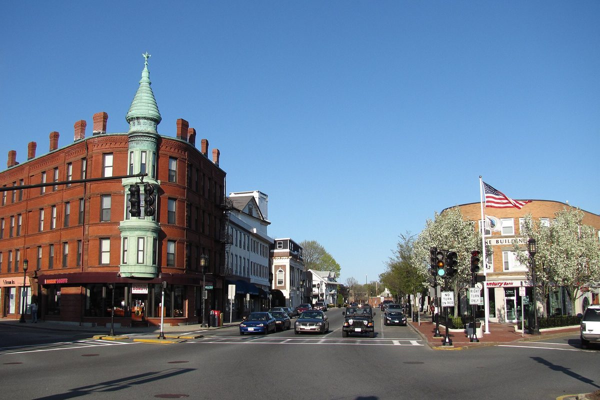 Medford Square: a red-brick corner block with a green copper turret, cars waiting at the light and an American flag over the sidewalk on a clear day