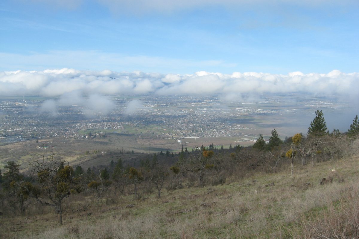 Medford and the Rogue Valley spread across the valley floor below a dry, grassy slope of Roxy Ann Peak, with a band of low cloud above