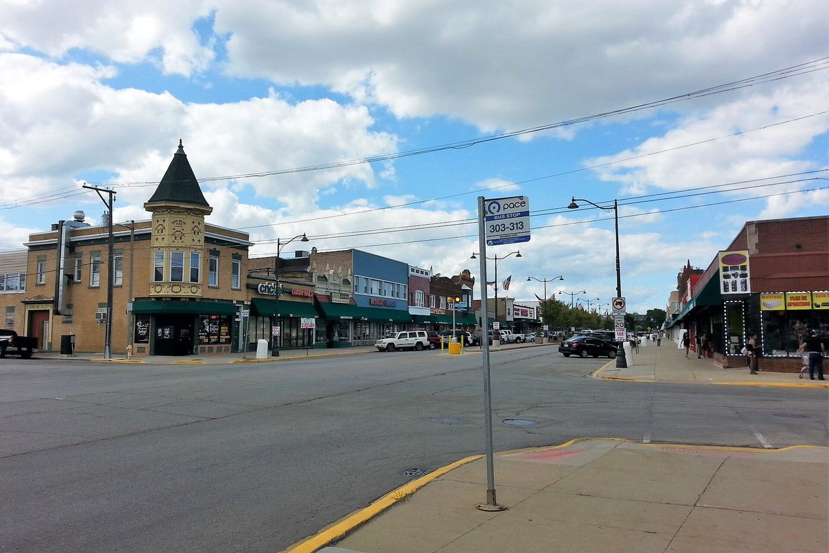 A downtown Melrose Park street corner with a tan brick building topped by a pointed turret, green shop awnings and a Pace bus stop sign under a cloudy blue sky