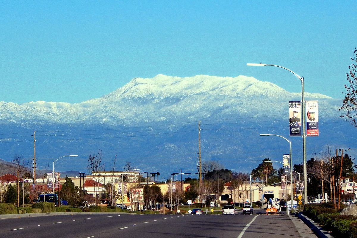 Snow-covered Mount San Jacinto rising behind Newport Road in Menifee, with streetlights, palm trees and low shops along the road