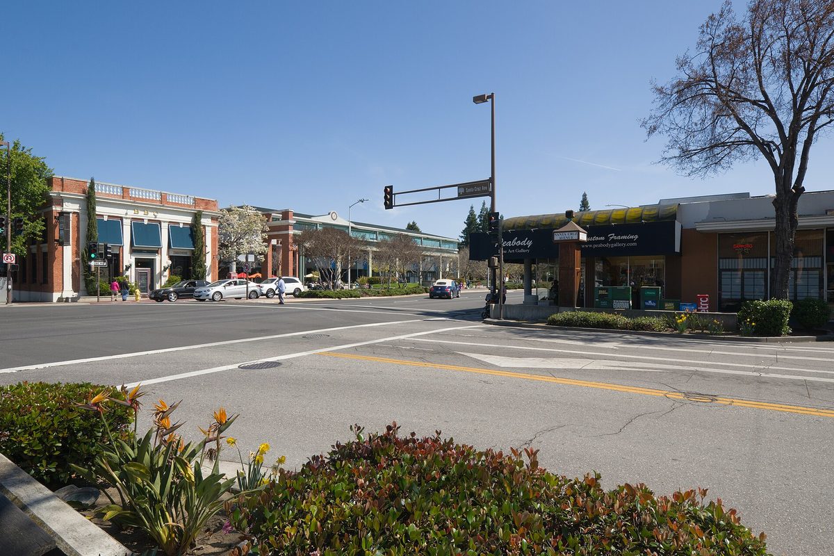 A wide downtown Menlo Park street with brick and glass storefronts, parked cars, a traffic signal and flowering shrubs in the foreground under a clear sky