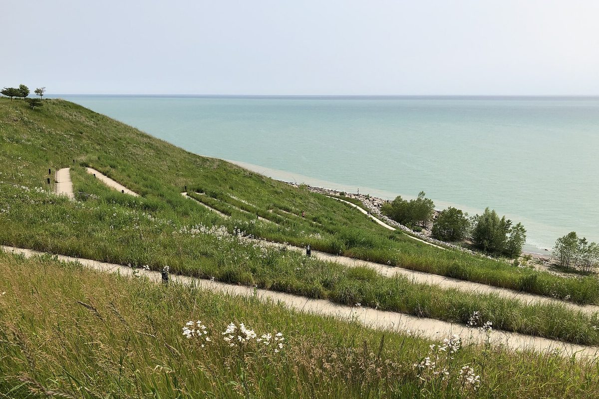 A grassy bluff with winding gravel paths and wildflowers sloping down to the calm, blue-green water of Lake Michigan in Mequon