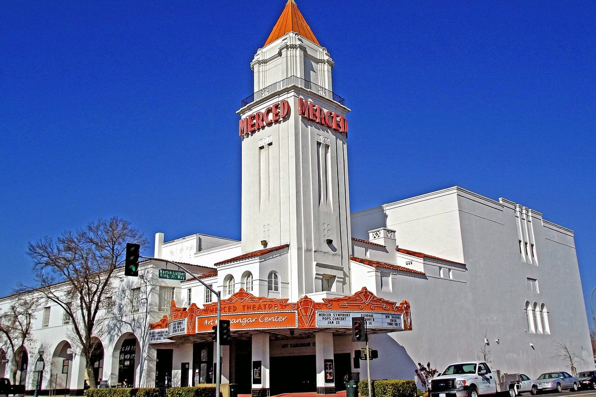The white Merced Theatre on Main Street, its tall tower lettered MERCED in red above an orange marquee, under a clear blue sky