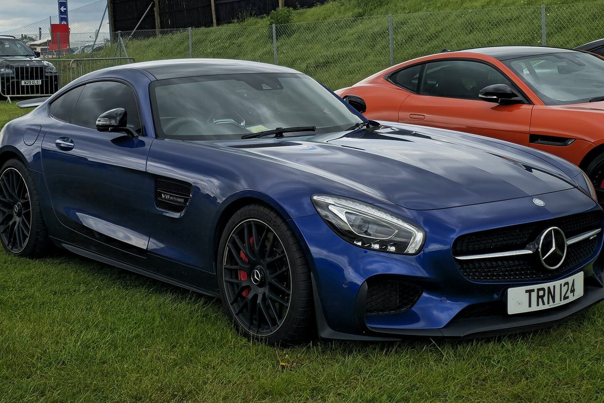 A dark blue Mercedes-AMG GT S coupe with black wheels and red brake calipers, parked on grass beside an orange sports car