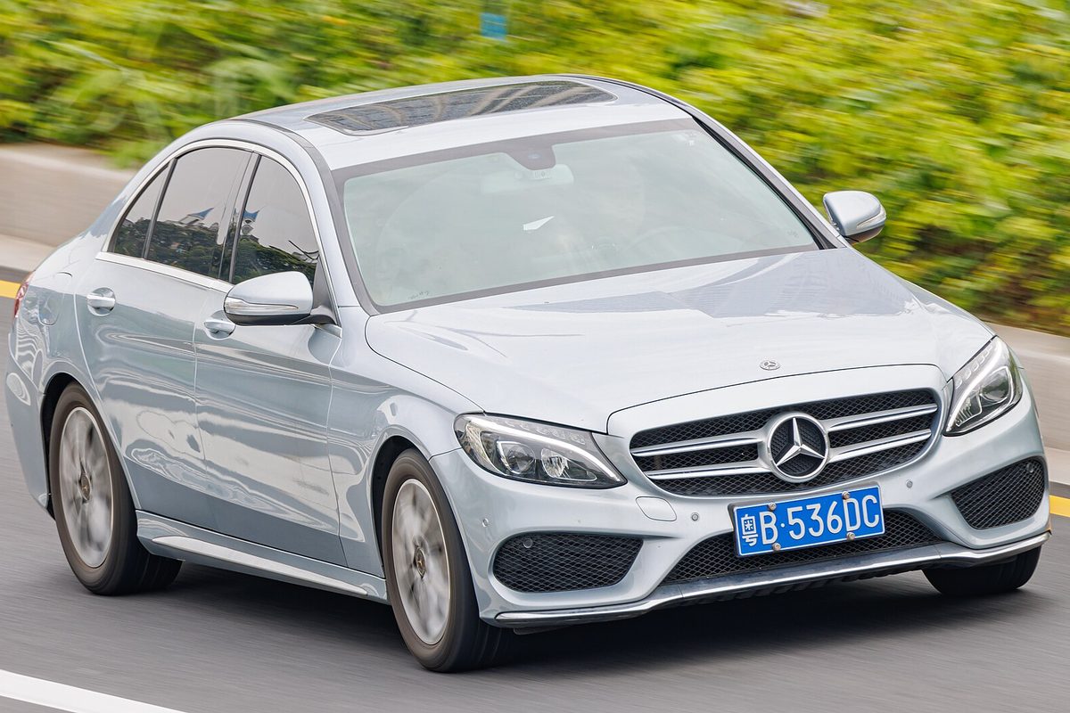 A light silver W205 Mercedes-Benz C-Class long-wheelbase sedan with a sunroof, driving past a green hedge