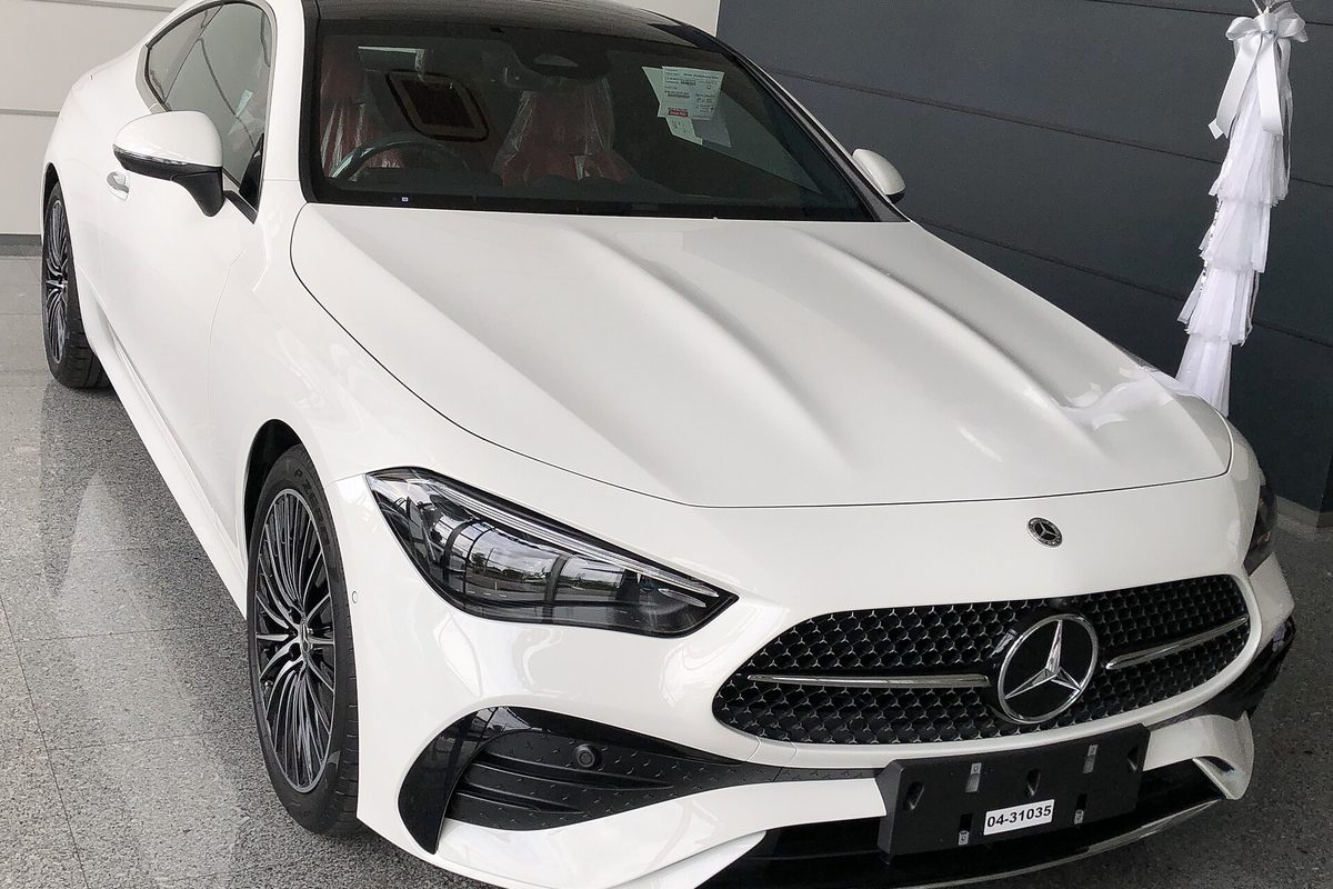 A white Mercedes-Benz CLE-Class coupe on a showroom floor, seen from the front, with a white ribbon decoration behind it
