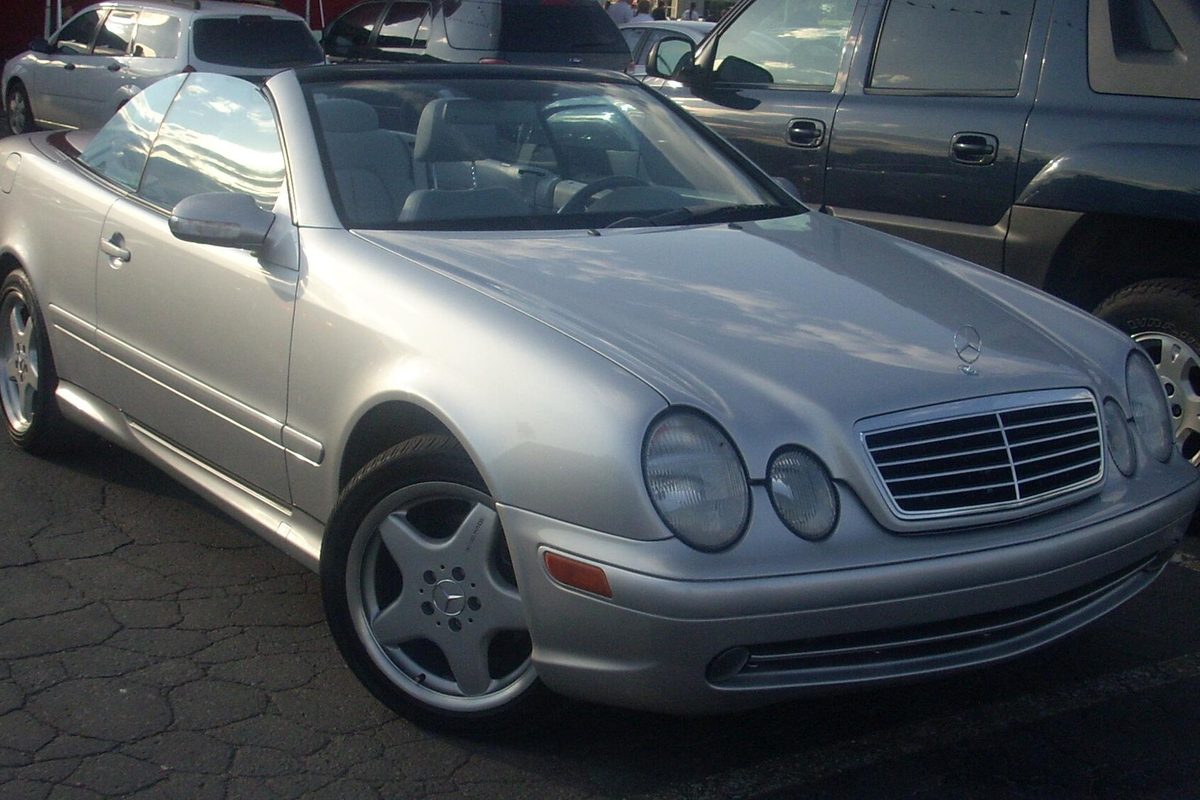 A silver first-generation Mercedes-Benz CLK convertible with the top down, parked in a lot beside an SUV