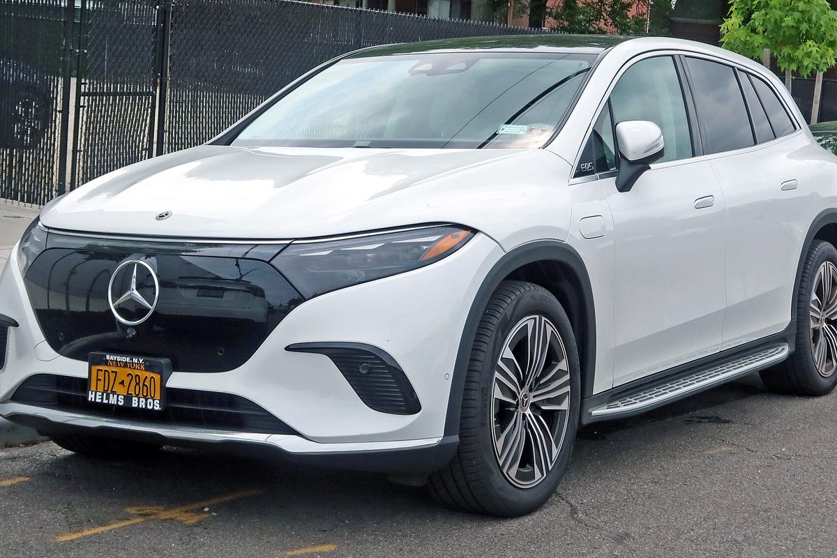 A white Mercedes-Benz EQS SUV parked on a city street in front of a chain-link fence