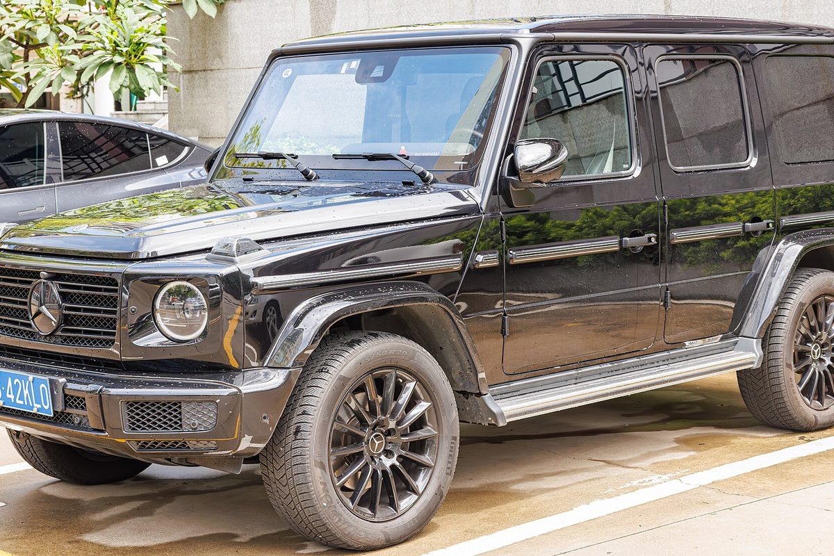 A black Mercedes-Benz G-Class SUV with dark multi-spoke wheels, parked on wet pavement beside another black car