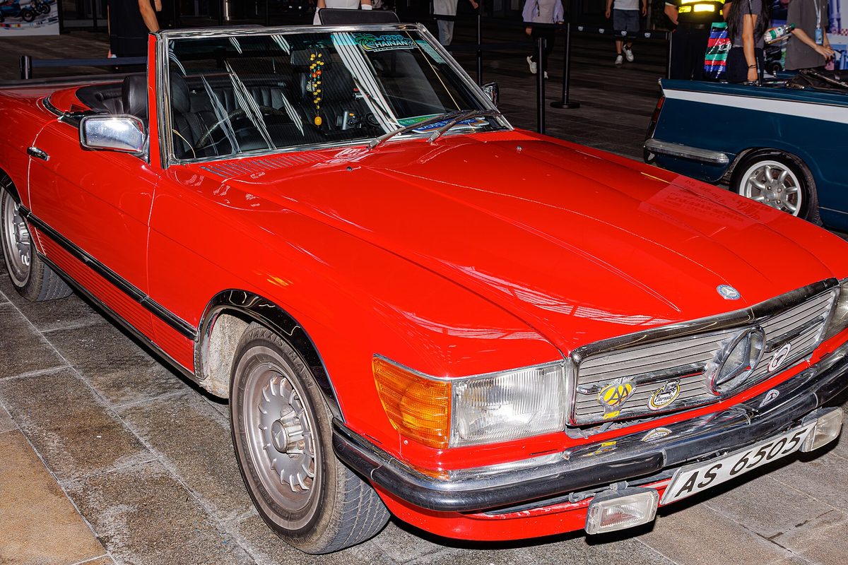A red R107 Mercedes-Benz SL convertible with the top down and chrome bumpers, parked on a paved plaza at night