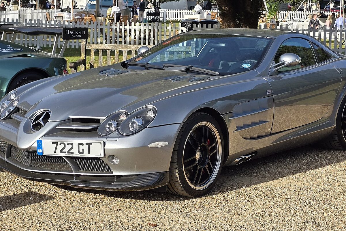 A silver Mercedes-Benz SLR McLaren coupe parked on gravel at a car show, a white picket fence and spectators behind it