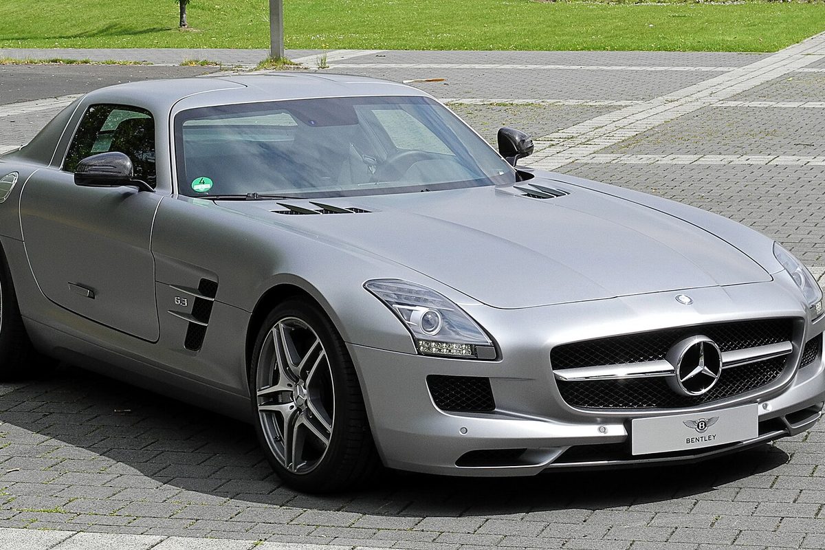 A silver Mercedes-Benz SLS AMG gull-wing coupe parked on a paved plaza, seen from the front corner