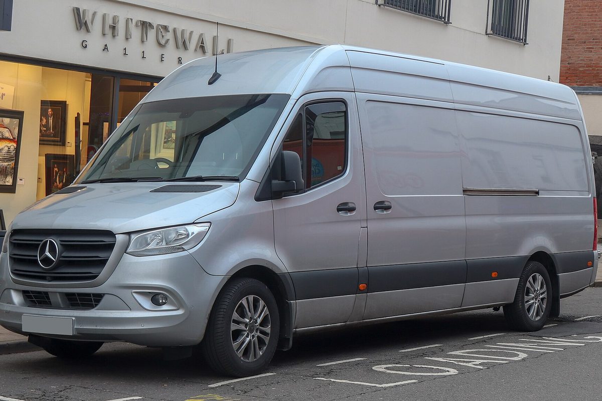 A silver high-roof Mercedes-Benz Sprinter panel van parked at a curb in front of a shop window