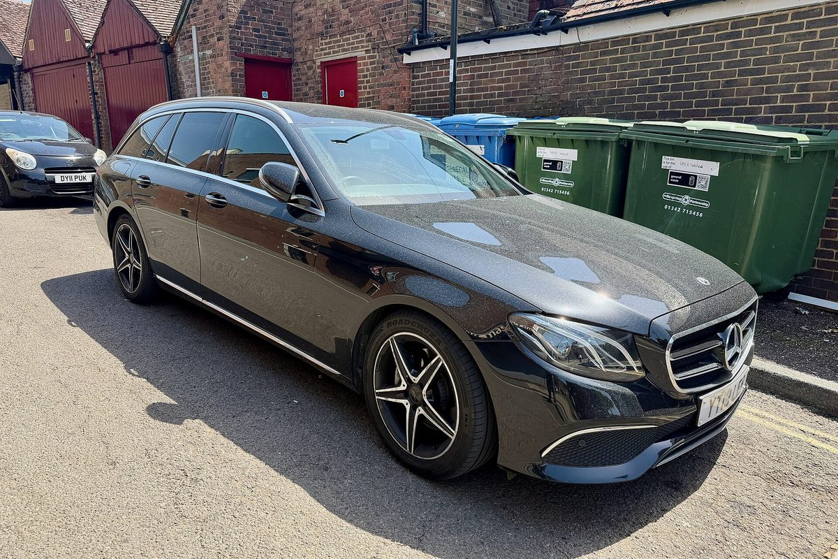 A black Mercedes-Benz E-Class estate wagon from the 2018 model year parked on a sunny lane beside green wheelie bins and a brick building