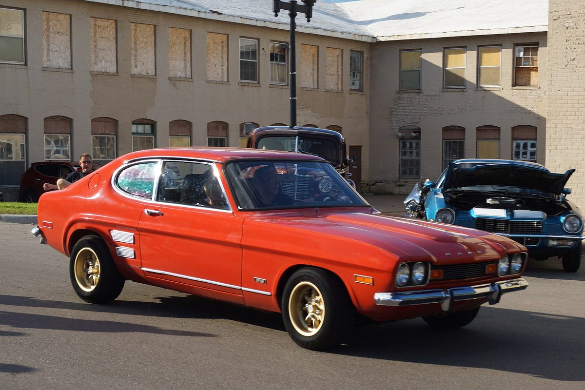 An orange Mercury Capri coupe with gold wheels and white side stripes, driving past old brick buildings at a car show