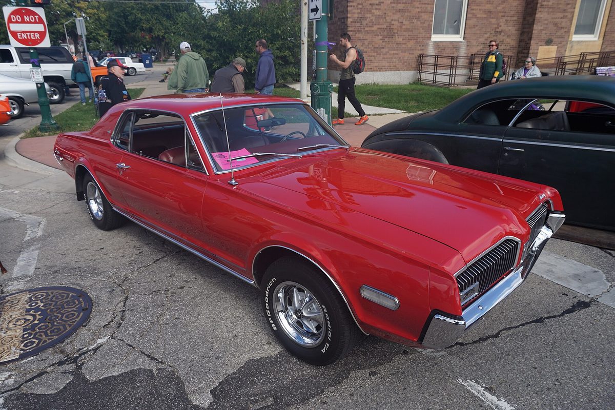 A red 1968 Mercury Cougar two-door hardtop with chrome bumpers at an outdoor classic car show, with spectators on the sidewalk behind it