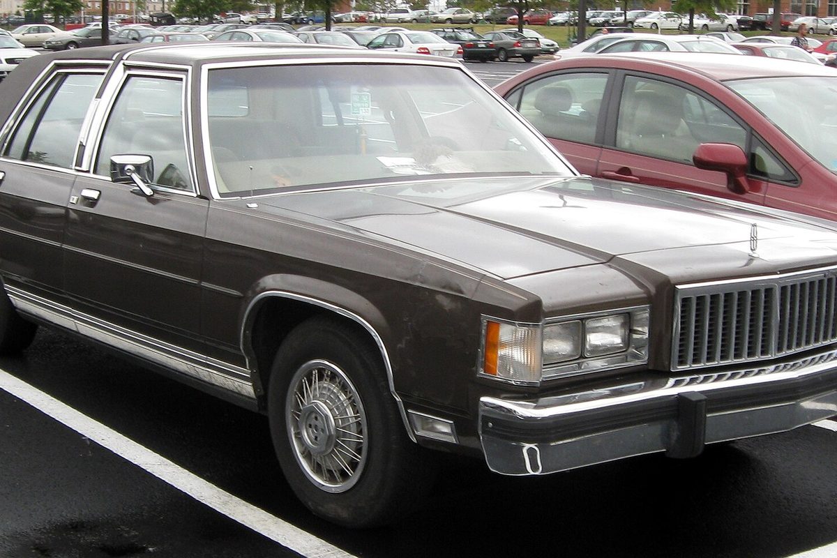 A dark brown 1983-1987 Mercury Grand Marquis four-door sedan with a chrome grille and wire-style hubcaps, parked in a busy lot
