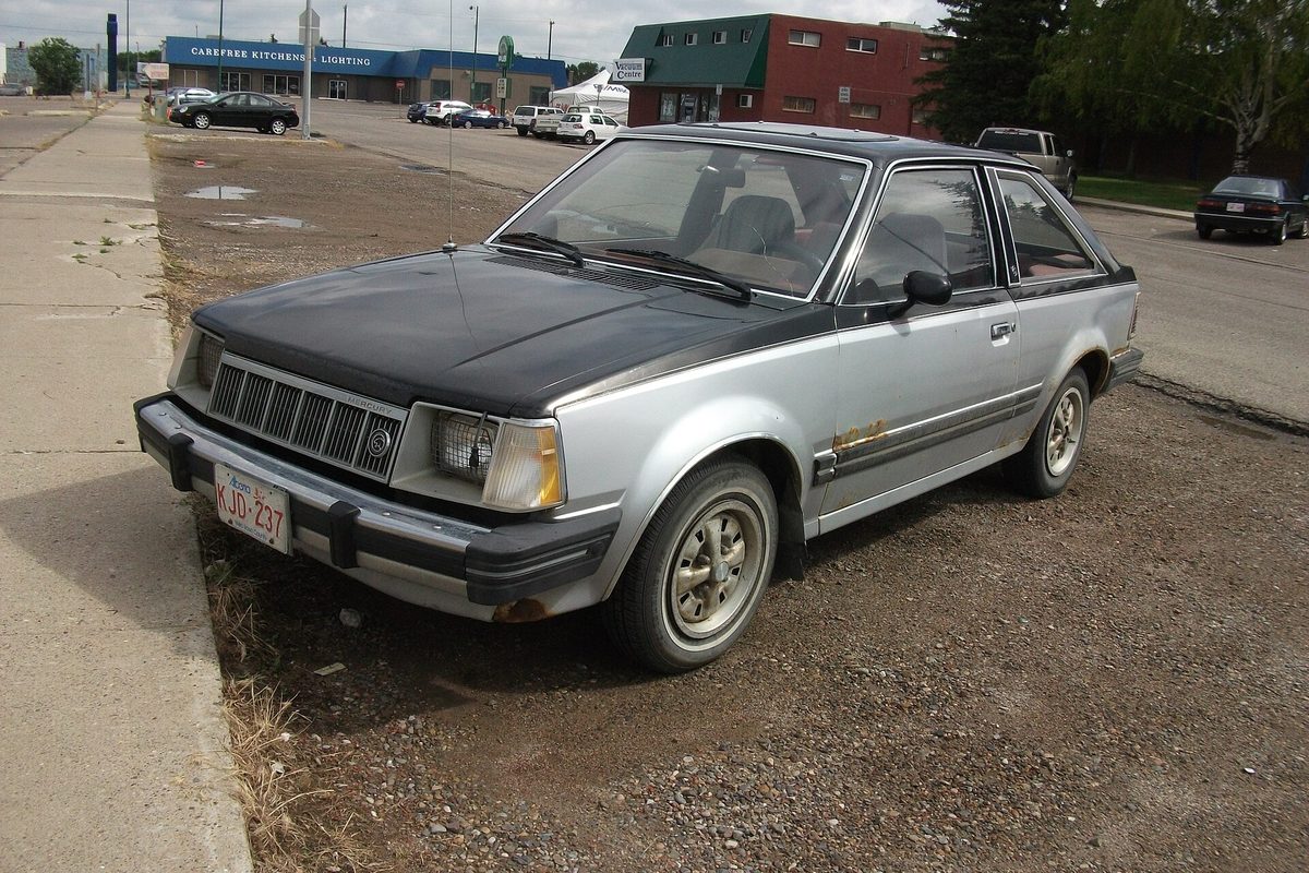 A silver Mercury Lynx three-door hatchback with a black hood, parked on dirt beside a sidewalk