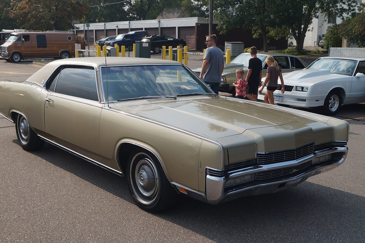 A gold 1970 Mercury Marquis Brougham two-door hardtop with a vinyl roof and hidden headlights, parked at a car show
