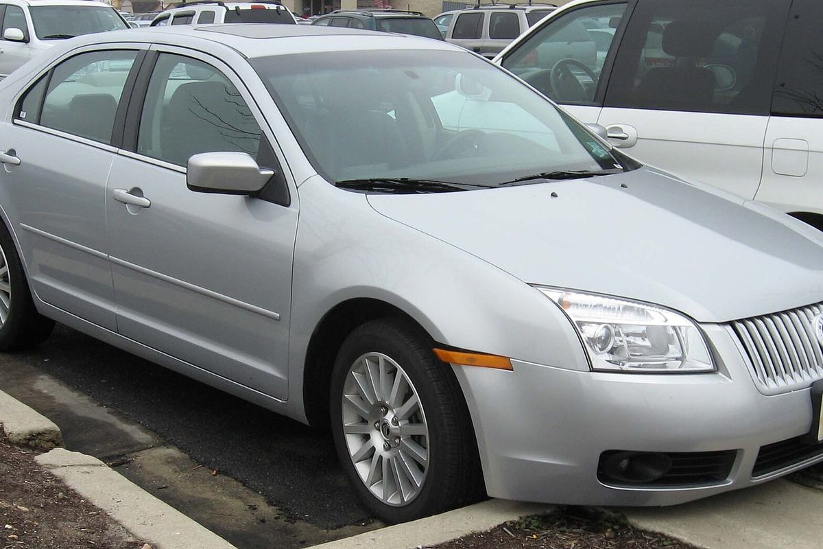 A silver 2006-2007 Mercury Milan sedan with a vertical-bar chrome grille, parked at a curb between other cars