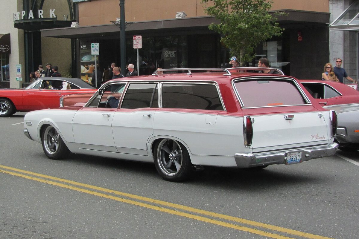 An early Mercury Montego station wagon, white with a red roof and a roof rack, rolling down a street past a car show crowd