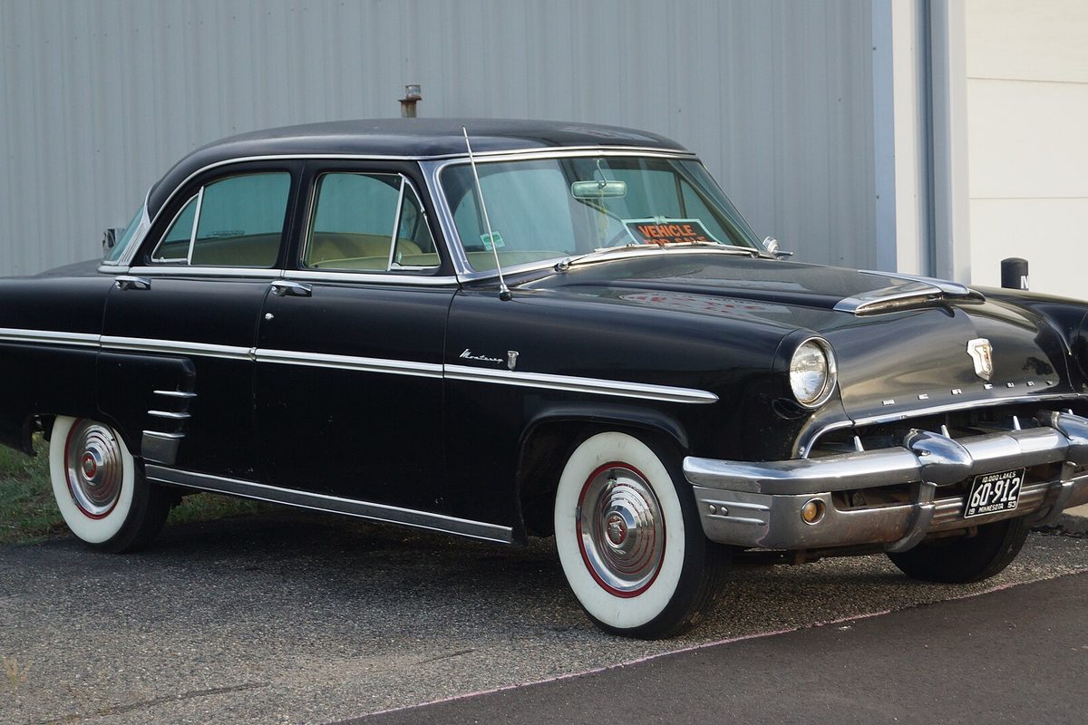 A black vintage Mercury Monterey four-door sedan with whitewall tires and chrome trim, parked beside a gray metal building