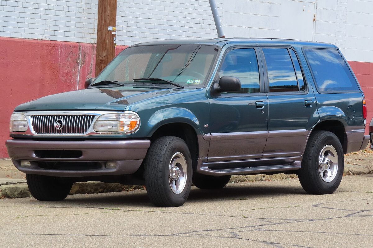 A green two-tone 1997 Mercury Mountaineer SUV with gray lower panels, parked on a cracked street beside a red and white painted brick wall