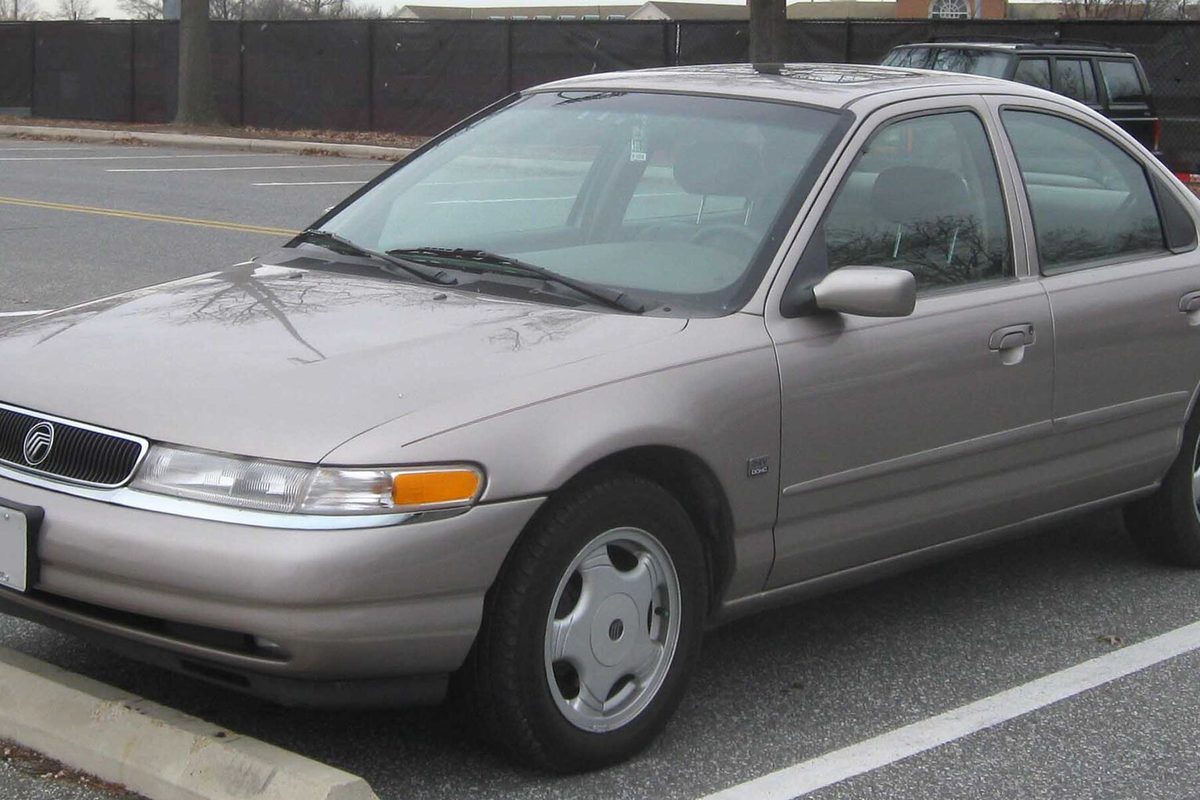 A taupe 1995-1997 Mercury Mystique four-door sedan with plastic wheel covers, parked in a lot with bare trees behind it
