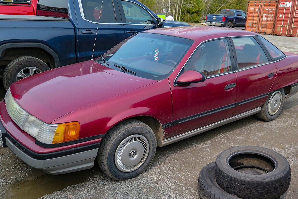 A dark red 1988 Mercury Sable GS sedan parked on gravel next to a stack of old tires, with a blue pickup behind it