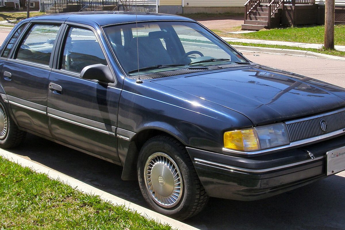 A dark blue 1988-1991 Mercury Topaz four-door sedan with wheel covers, parked at a curb next to a lawn