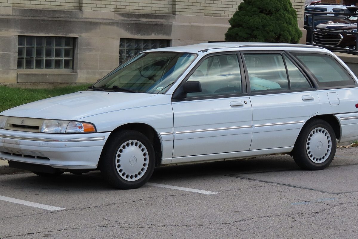 A white 1995 Mercury Tracer station wagon with white wheel covers, parked at a curb beside a stone building