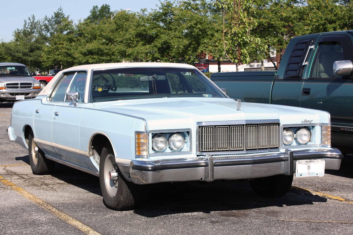 A pale blue 1970s Mercury Grand Marquis four-door with a chrome upright grille and twin round headlights, parked in a lot next to a green pickup