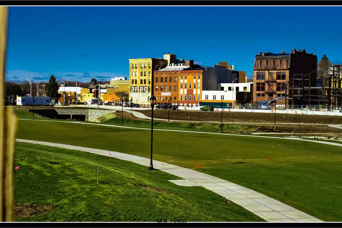 Downtown Meriden's old brick and painted commercial buildings seen across the new green lawn and curving paths of the park at the HUB, with black lampposts under a blue sky