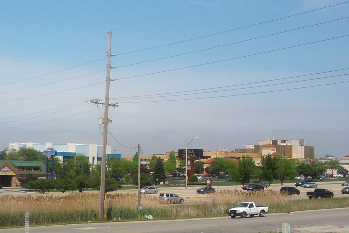 Merrillville seen across tall dry reeds beside US 30, with a white pickup on the road and low commercial buildings and a tall sign behind