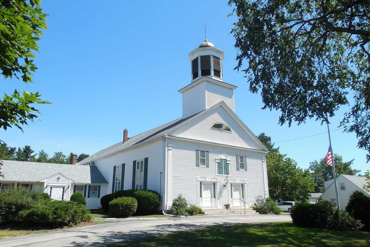 The white clapboard First Congregational Church in Merrimack, with a domed cupola, green shutters and an American flag on a pole beside it