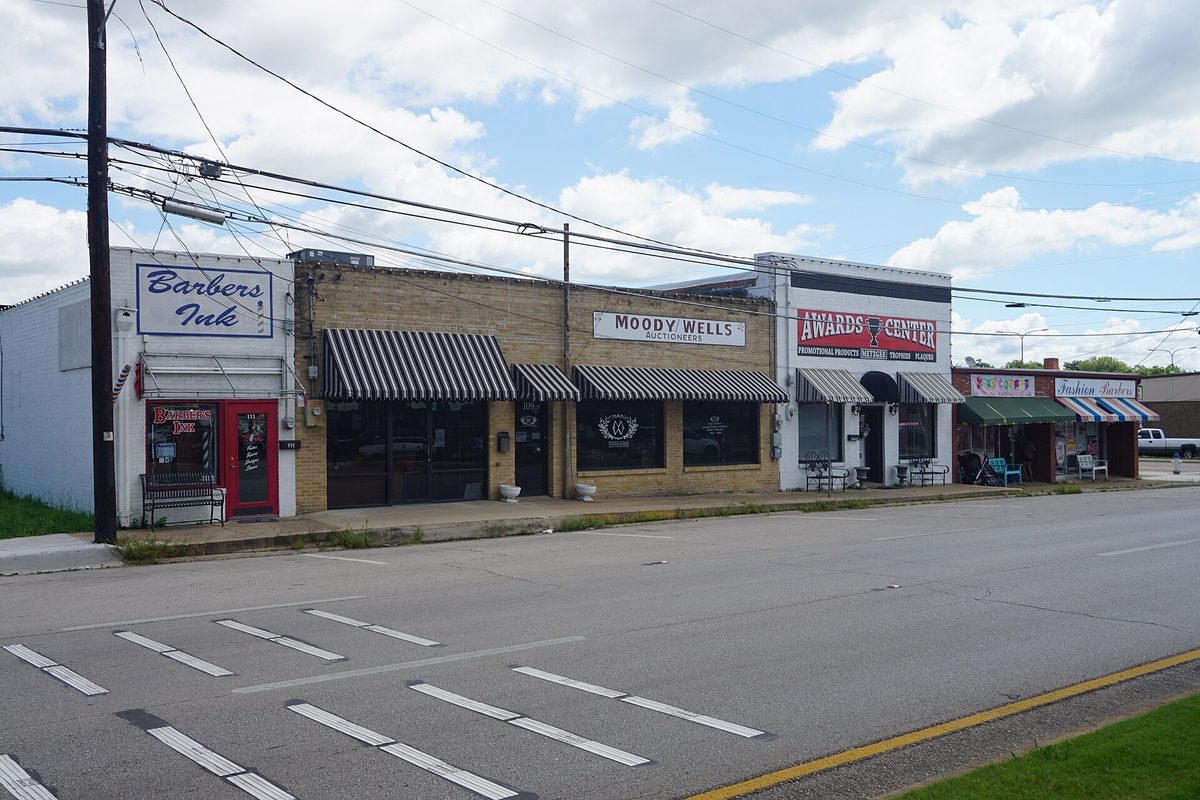 A row of small storefronts with striped awnings on Main Street in Mesquite, including a barber shop and an awards shop, under power lines