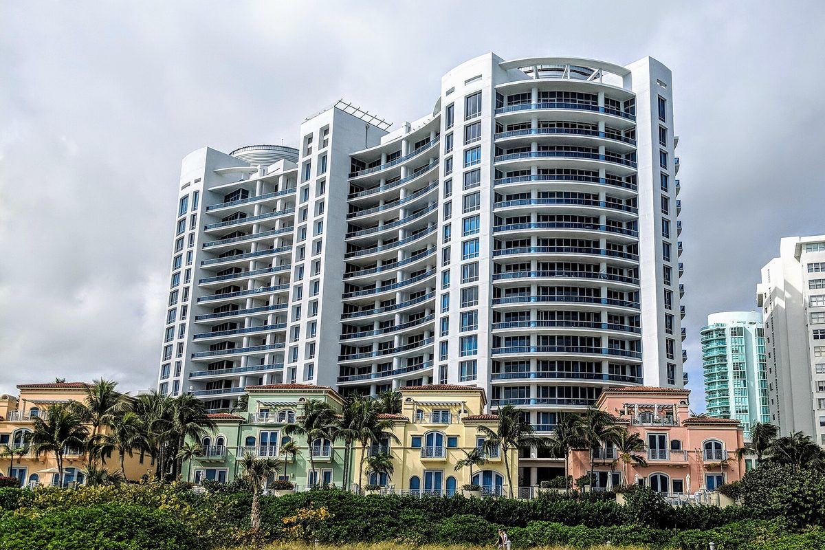 White curved condominium towers rising behind pastel low-rise buildings, palm trees and dense green shrubs in Miami Beach under a gray sky