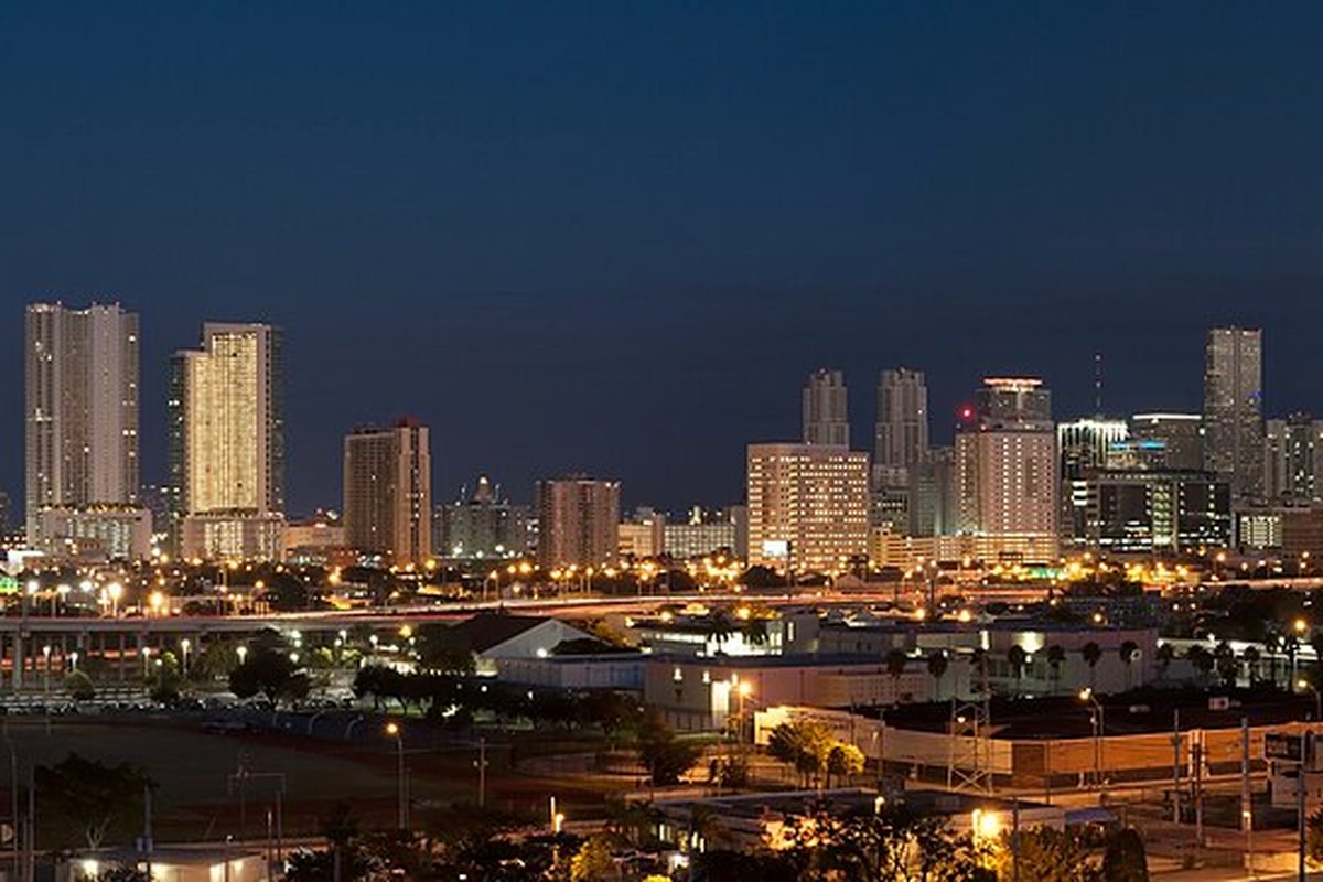 Downtown Miami skyline at dusk, lit high-rise towers above a raised highway and low warehouses in the foreground