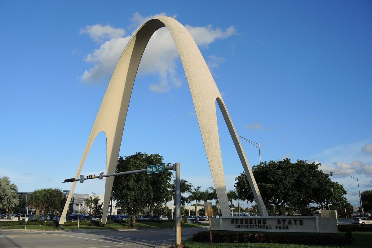 The tall white Sunshine State Arch over the entrance sign of Sunshine State International Park, next to a NW 167 St street sign and traffic light