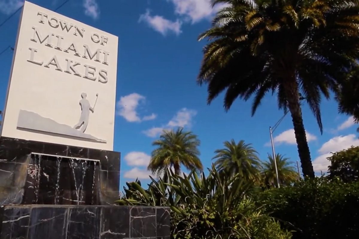The stone Town of Miami Lakes entrance sign above a small water feature, with palm trees and a blue sky behind it