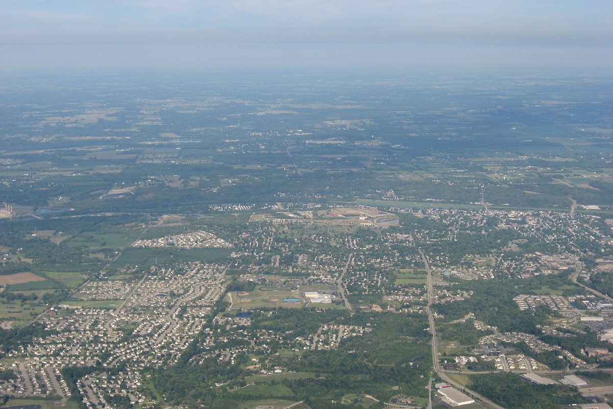 Hazy aerial view over Miamisburg from the east, with neighborhoods, roads and open fields in the foreground and flat farmland running to the horizon