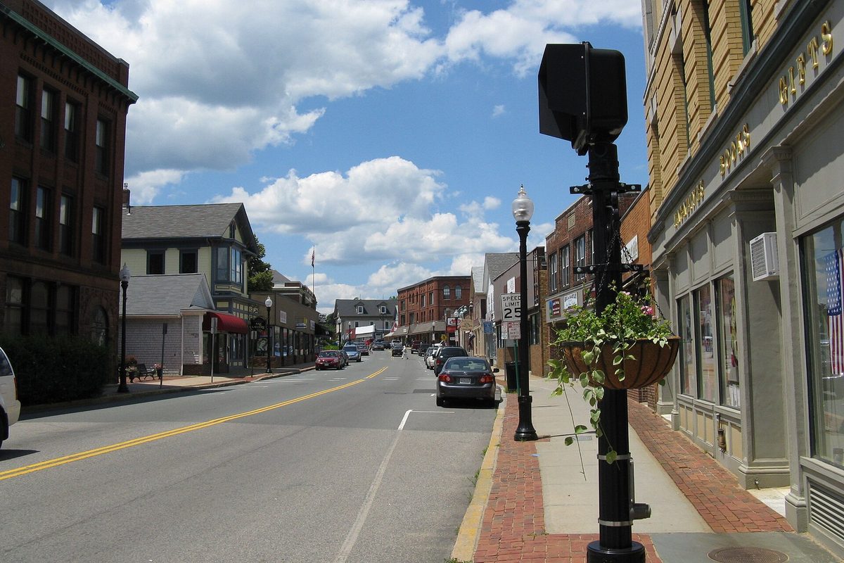 Centre Street in downtown Middleborough, lined with brick storefronts and lamp posts, with a hanging flower basket in the foreground on a sunny day