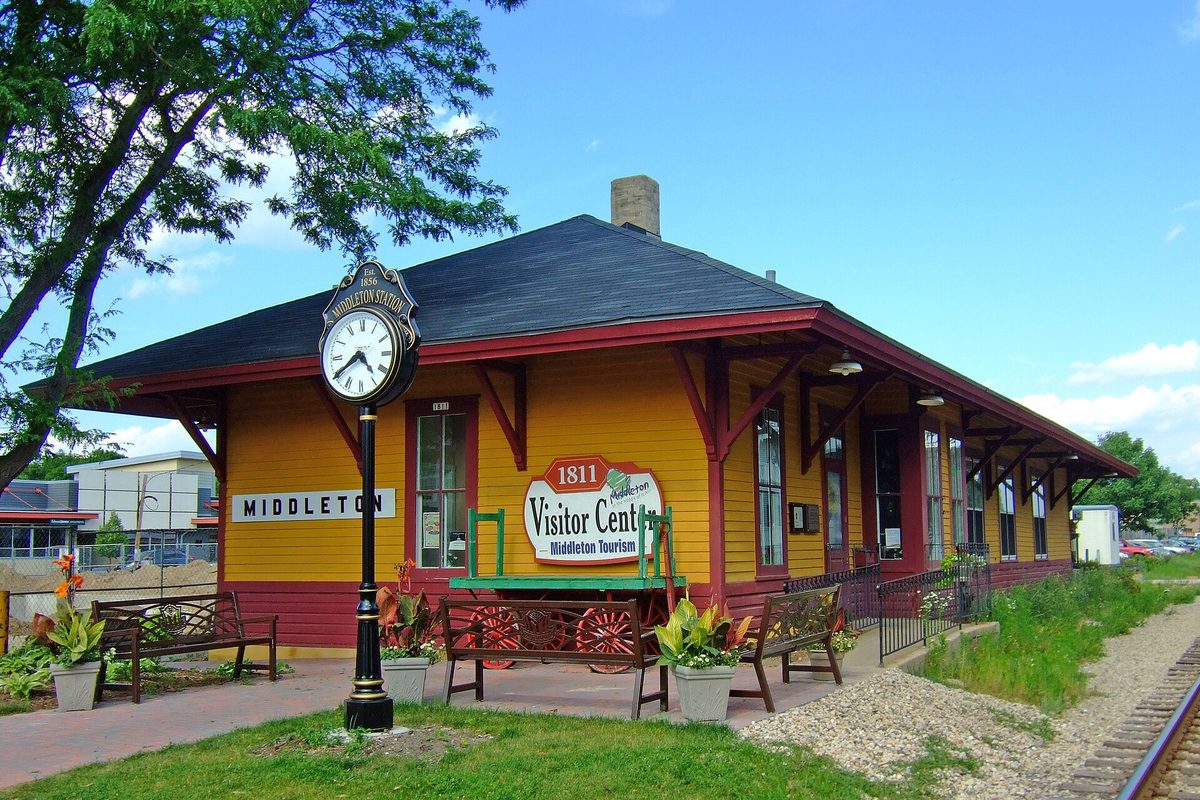 The yellow depot building with red trim that houses the Middleton Visitor Center, with a post clock, benches and railroad tracks running alongside